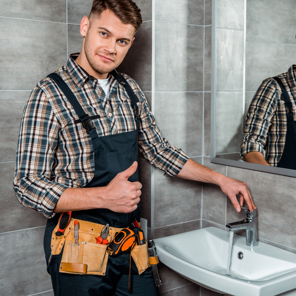 handsome installer standing near faucet with flowing water and showing thumb up - Fotó, kép