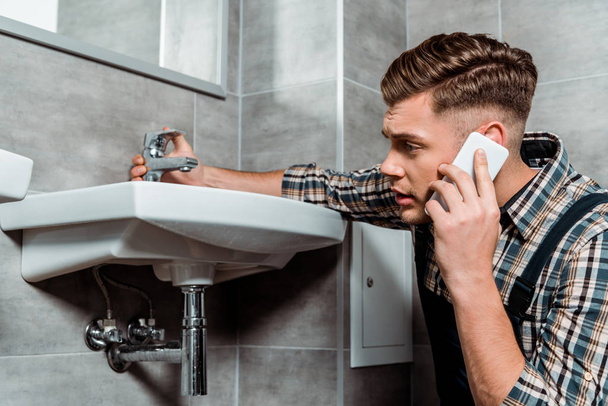 handsome installer touching faucet and talking on smartphone  - Photo, Image