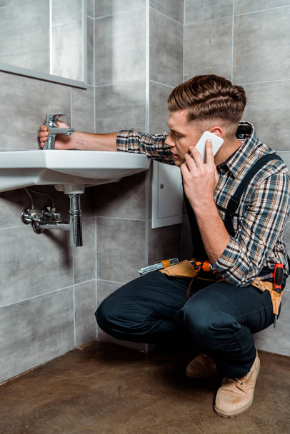 handsome installer sitting near sink and touching faucet while talking on smartphone  - Фото, зображення