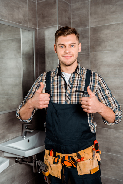 χαρούμενος εγκαταστάτης με ζώνη εργαλείων στέκεται στο μπάνιο και δείχνει τους αντίχειρες επάνω  - Φωτογραφία, εικόνα