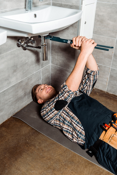 Glücklicher Installateur hält Gleitgelenkzange in Rohrnähe, während er im Badezimmer auf dem Boden liegt  - Foto, Bild