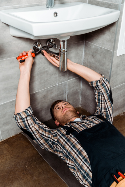 handsome installer holding pliers while lying on floor in bathroom  - Zdjęcie, obraz
