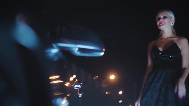 Piękna Blondynka pozowanie w czarnej sukience w nocy w pobliżu samochodu - Materiał filmowy, wideo