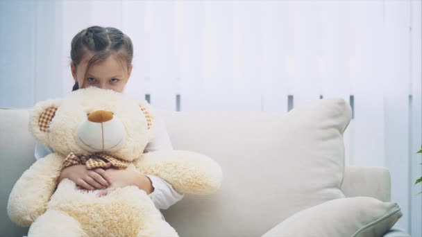 Happy little girl sitting on the sofa, dipping her head in the big teddy-bear, then looking up at the camera smiling. - Video