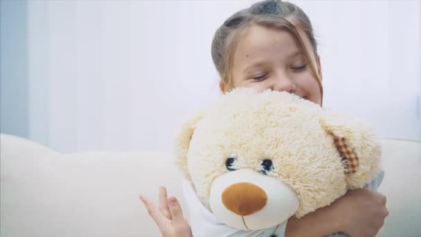 Happy little girl with plaits sitting on the sofa, hugging closely a big white teddy. - Imágenes, Vídeo