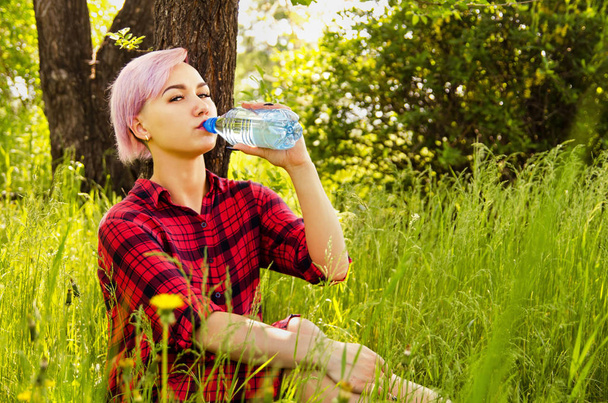 緑の草に座っている若い美しい女性は、木や茂みの背景に水のボトルを保持し、飲みます - 写真・画像