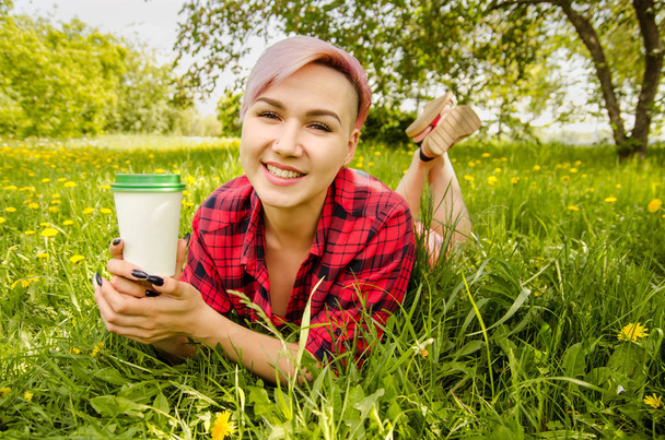 Young beautiful smiling girl lies on a green grass and dandelions and holds paper cup with coffee. - Foto, Bild