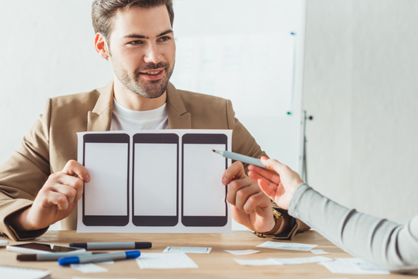 Выборочный фокус улыбающегося дизайнера планирования пользовательского опыта проектирования мобильного макета с коллегой в офисе
 - Фото, изображение