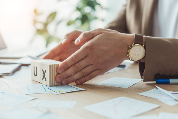 Oříznutý pohled návrháře držícího dřevěné kostky s písmeny ux vedle náčrtků aplikací na stole - Fotografie, Obrázek