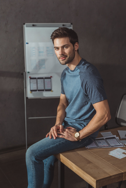 Handsome designer looking at camera while sitting on table with layouts  - Photo, Image