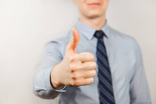 Young businessman dressed in blue shirt and tie shows thumb up close up, on gray background - Foto, Imagen
