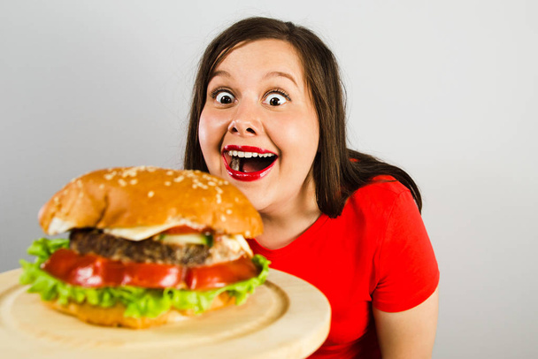 Young fat woman holds hamburger, looks at him and smiles on light pink background. - Foto, Imagem