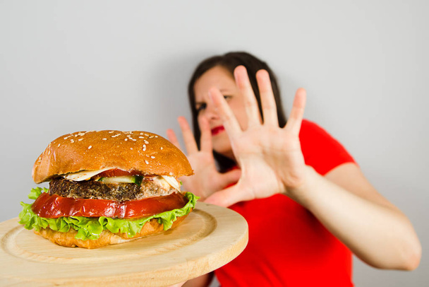 Young girl refuses fat unhealthy hamburger because she is losing weight and dieting on gray background. - Foto, Imagem