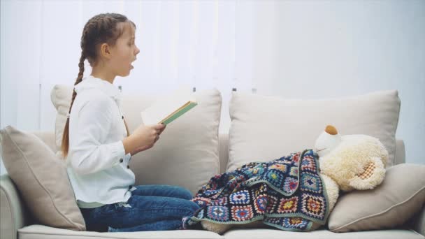 Cute little girl sitting with her legs curled up, reading fairy-tale for her teddy, that is lying under the blanket. - Imágenes, Vídeo