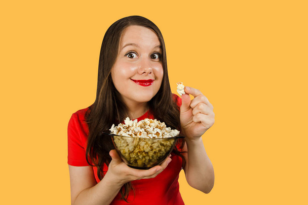 Young pretty girl with opened mouth and wide eyes holds glass bowl with popcorn isolated on orange background - Foto, Bild
