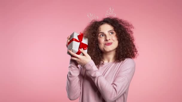 Young cute girl smiling and holding present box on pink studio background. Woman in Christmas head horns deer wreath. New year mood - Кадры, видео