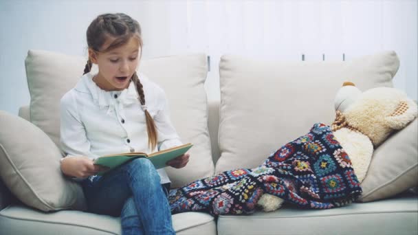 Cute little girl sitting on the sofa, reading fairy-tale for her teddy, that is lying under the blanket. - Footage, Video