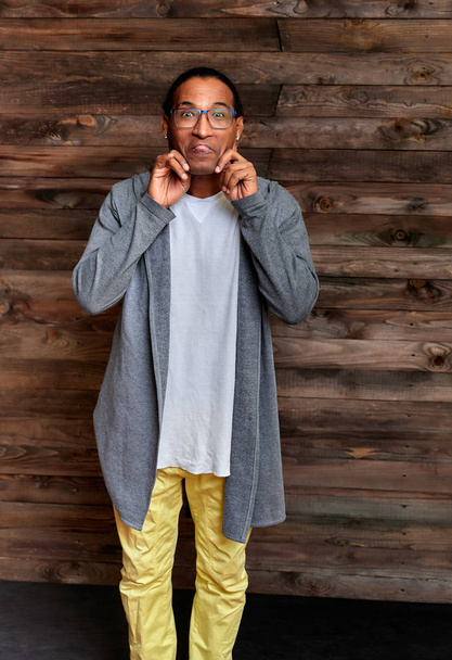 Vertical Portrait Young man with short-haired African-American glasses in ordinary clothes on a vintage wooden background in a home interior in a loft style. Standing right in front of the camera - Photo, image