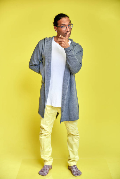 Vertical portrait of a cheerful young man wearing glasses African-American with a short haircut in ordinary clothes on a yellow background in full length. Standing right in front of the camera - 写真・画像