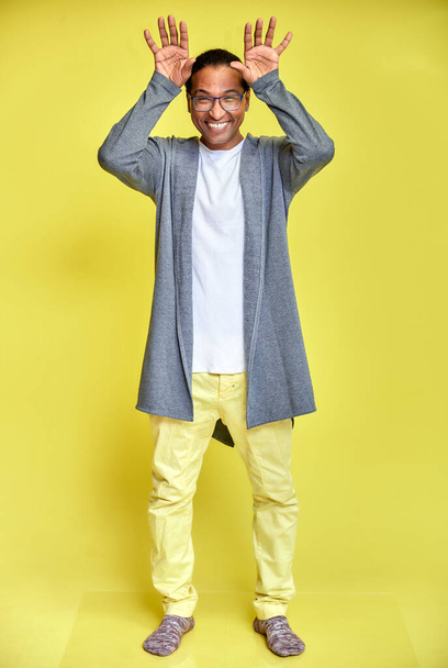 Vertical portrait of a cheerful young man wearing glasses African-American with a short haircut in ordinary clothes on a yellow background in full length. Standing right in front of the camera - Foto, Bild