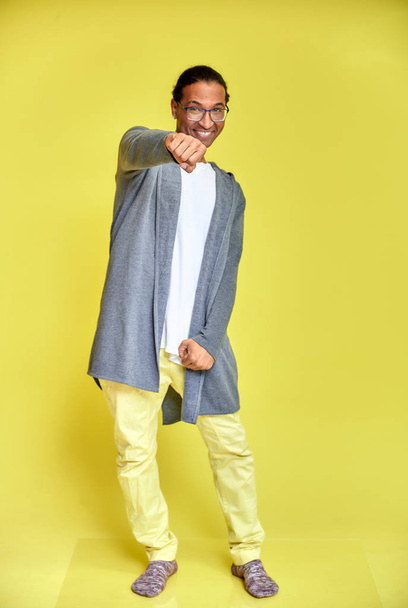 Retrato vertical de um jovem alegre usando óculos afro-americanos com um corte de cabelo curto em roupas comuns em um fundo amarelo de comprimento total. Mesmo em frente à câmara
 - Foto, Imagem