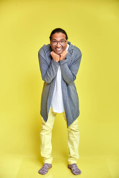 Vertical portrait of a cheerful young man wearing glasses African-American with a short haircut in ordinary clothes on a yellow background in full length. Standing right in front of the camera - Foto, Imagem