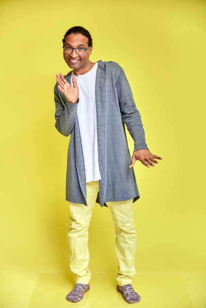 Vertical portrait of a cheerful young man wearing glasses African-American with a short haircut in ordinary clothes on a yellow background in full length. Standing right in front of the camera - Fotografie, Obrázek