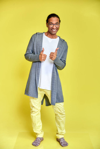 Vertical portrait of a cheerful young man wearing glasses African-American with a short haircut in ordinary clothes on a yellow background in full length. Standing right in front of the camera - Zdjęcie, obraz