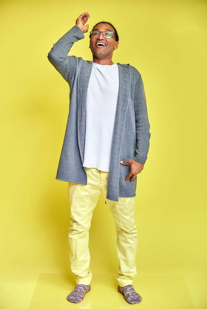 Vertical portrait of a cheerful young man wearing glasses African-American with a short haircut in ordinary clothes on a yellow background in full length. Standing right in front of the camera - Foto, Imagen