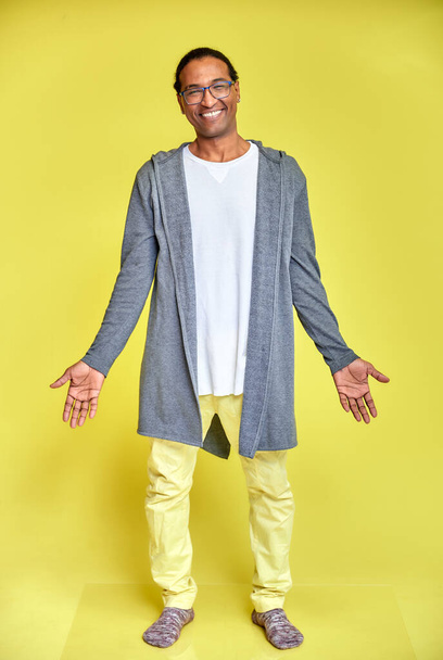 Vertical portrait of a cheerful young man wearing glasses African-American with a short haircut in ordinary clothes on a yellow background in full length. Standing right in front of the camera - Valokuva, kuva