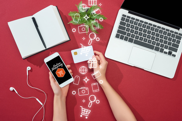 cropped view of woman holding credit card and smartphone with online shopping illustration near laptop, earphones, pen, notebook and plant on red background - Foto, Imagem