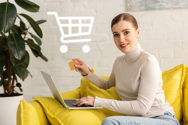 Beautiful girl with credit card using laptop and smiling at camera near cart illustration - Photo, Image