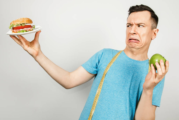 Young smiling guy with a measuring tape hanging on the shoulders, holds a burger and green apple, on a white background. Concept of choosing between healthy and unhealthy foods - Foto, imagen