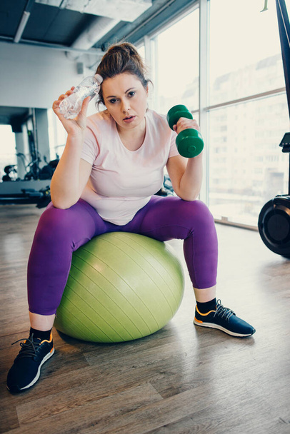 Tired fat woman exercises with dumbbells while sitting on gym ball in gym - Foto, immagini