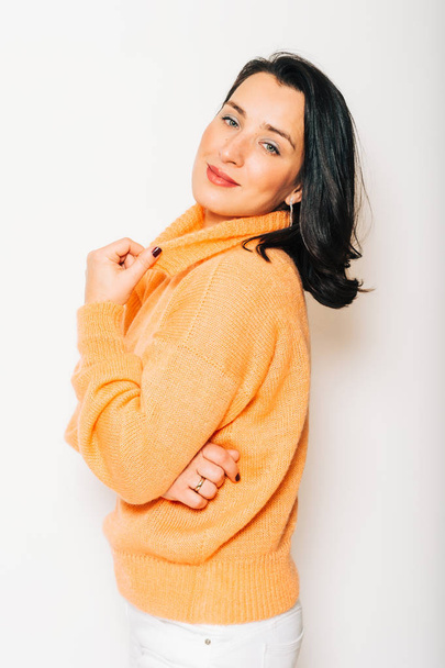 Studio portrait of beautiful brunette woman wearing orange pullover, posing on white background - Фото, изображение