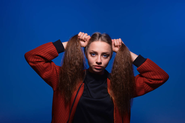 Cute teenager girl making ponytails holding hair with hands, posing in studio with blue background. - Foto, Imagen