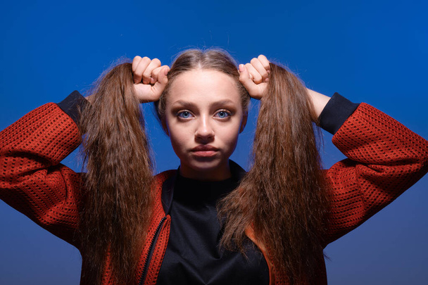 Cute teenager girl making ponytails holding hair with hands, posing in studio with blue background. - Fotografie, Obrázek