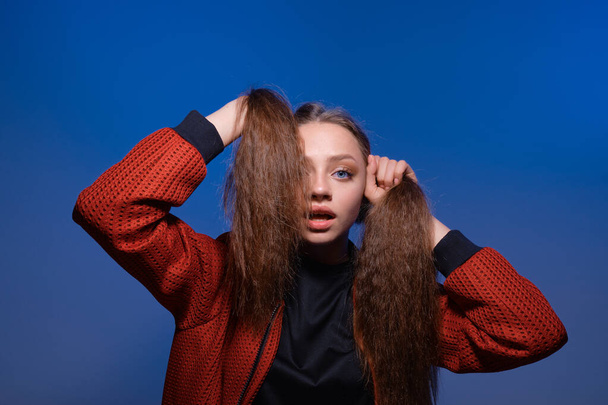 Cute teenager girl making ponytails holding hair with hands, posing in studio with blue background. - 写真・画像