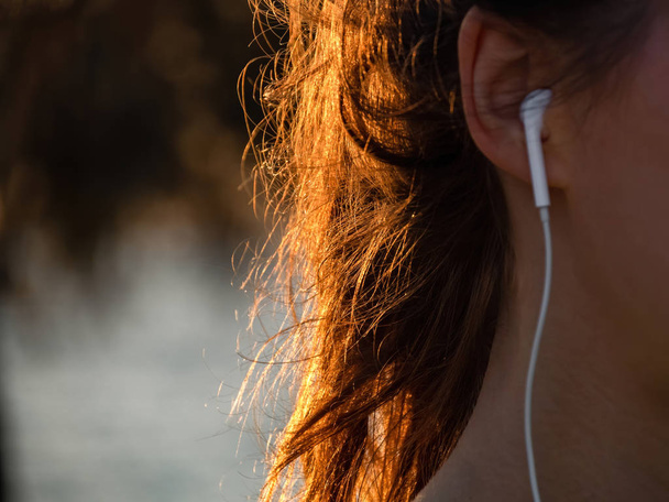 Een meisje met mooi zonlicht in het haar luistert naar muziek met oortjes. Geen gezicht.. - Foto, afbeelding