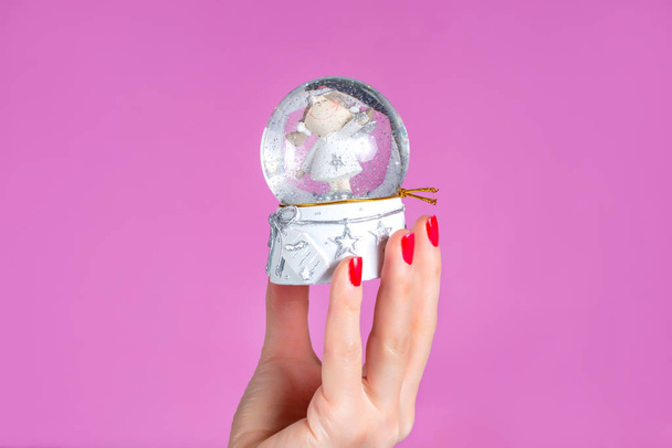 Mujer mano sosteniendo bola de adorno con nieve aislada sobre fondo rosa en el estudio. Manos femeninas con esmalte de uñas rojo en los dedos. Navidad o San Valentín vacaciones y el concepto de minimalismo
 - Foto, imagen