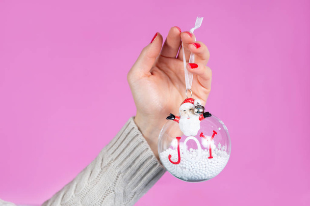 Vrouwelijke hand houden ornament bal met sneeuw geïsoleerd op roze achtergrond in de studio. Vrouwelijke handen met rode nagels polijsten op vingers. Kerstmis of Valentijnsdag vakantie en minimalisme concept - Foto, afbeelding