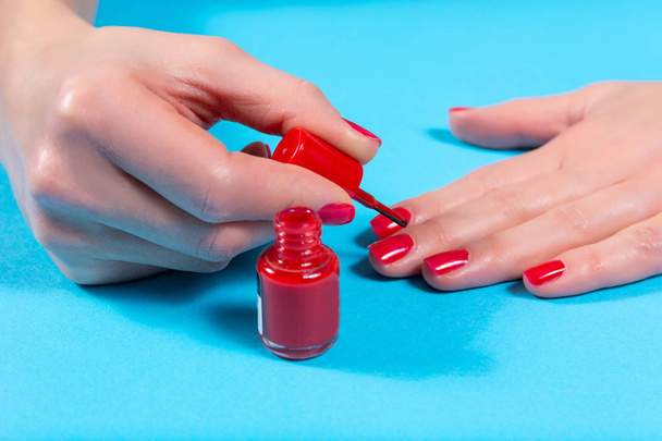 Mädchen, das rote Nagellackfarbe mit einem Pinsel auf der Hand aufträgt, isoliert auf blauem Hintergrund im Studio. Maniküre und Nagellack Konzept. Nahaufnahme, selektiver Fokus - Foto, Bild