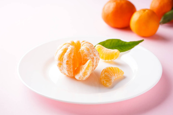 Tangerines with a green branch and slices in a white plate on a pink background.  - Zdjęcie, obraz