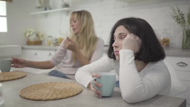 Close-up of adult brunette Caucasian woman siting with coffee cup at the table. Her blond female friend drinking hot drink at the background. Upset lady spending day with fellow. Friendship, resting. - Felvétel, videó