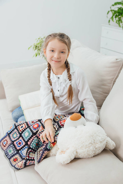 Smiling little girl with nice plaits is sitting on the sofa, near her teddy-bear, that is lying under the blanket. - Foto, afbeelding