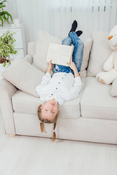 Funny little kid is lying on the sofa upside down, showing tongue, legs on the back of the sofa, plaits hang in the air, book in her hands, teddy bear is on the side. - Фото, изображение