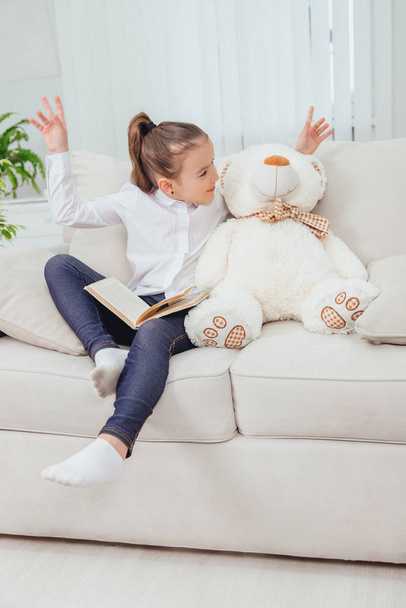 Cute little girl sitting on the sofa with teddy-bear near her, reading fairy-tale. - Foto, Imagem