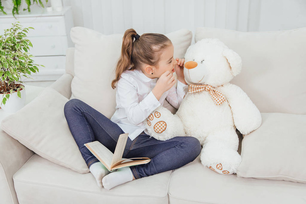 Cute little girl sitting on the sofa with teddy-bear near her, reading fairy-tale, setting her teeeth, imitating monster with arrrrr sound. - Foto, Imagen