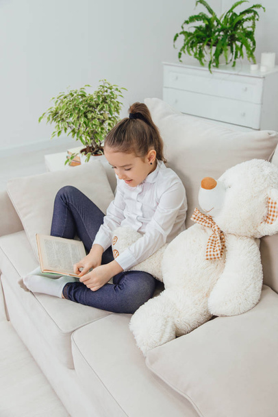 Cute little girl sitting on the sofa with teddy-bear near her, reading fairy-tale. - Фото, изображение