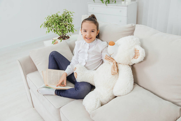 Cute little girl sitting on the sofa with teddy-bear near her, reading fairy-tale, pointing her finger at something in the book, smiling. - Foto, imagen
